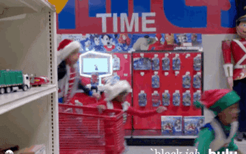 A Santa Clause and kids dressed as elves run through a store and throw toys into a shopping cart.