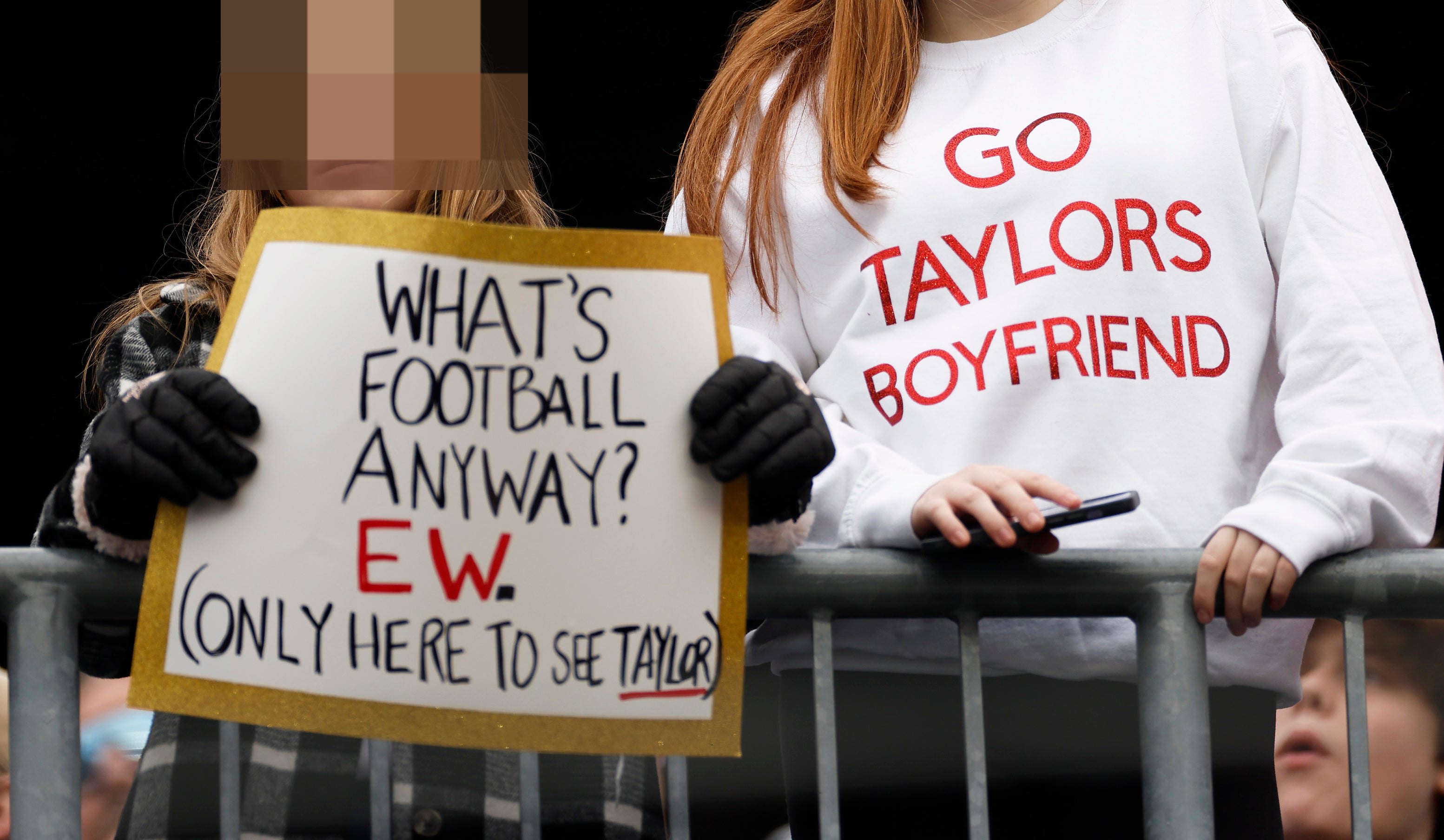 Close-up of someone holding the sign &quot;What&#x27;s football anyway? EW (only here to see Taylor)&quot; and someone wearing a &quot;Go Taylors boyfriend&quot; sweatshirt