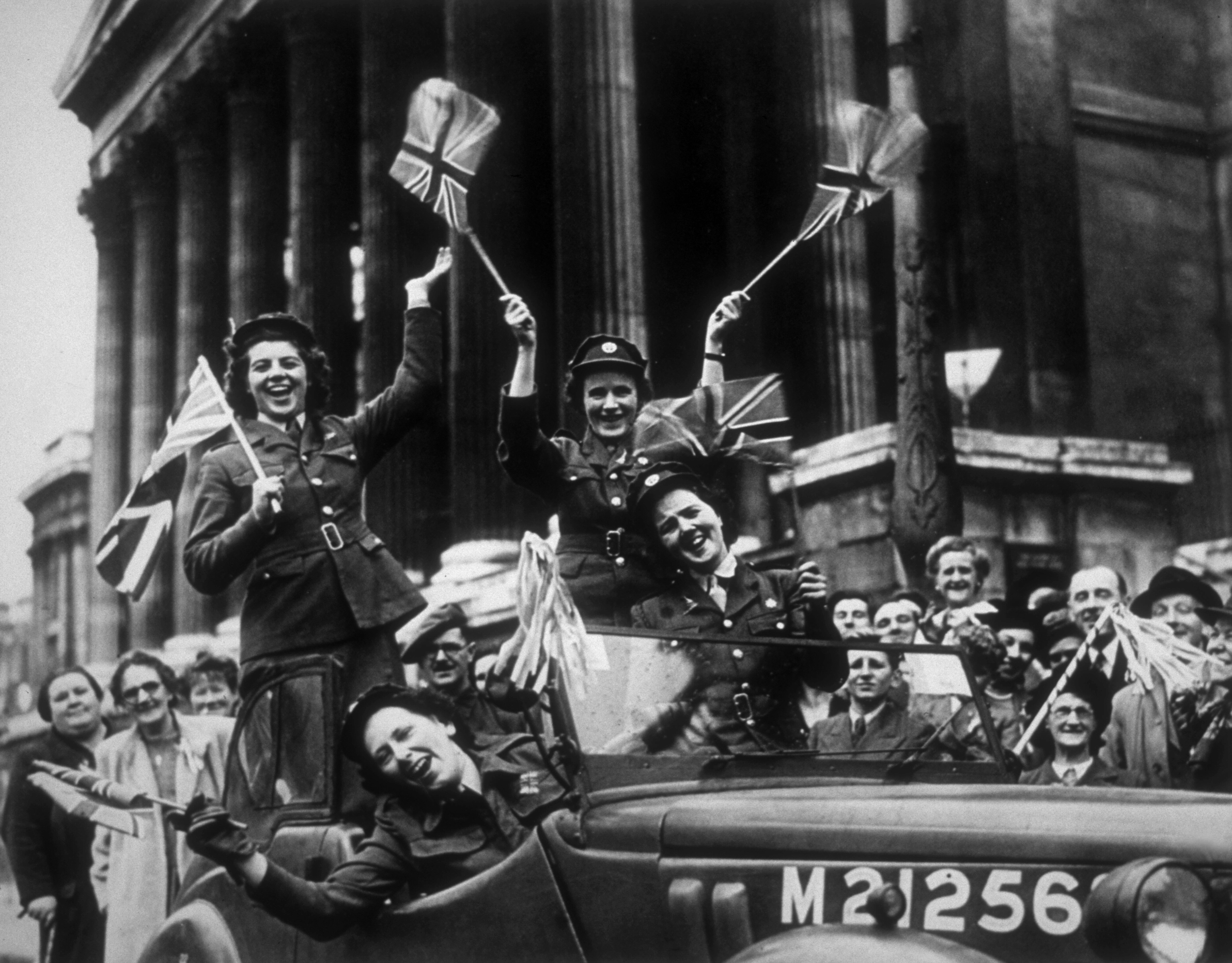 An old photograph of women in uniform in a car waving UK flags and smiling