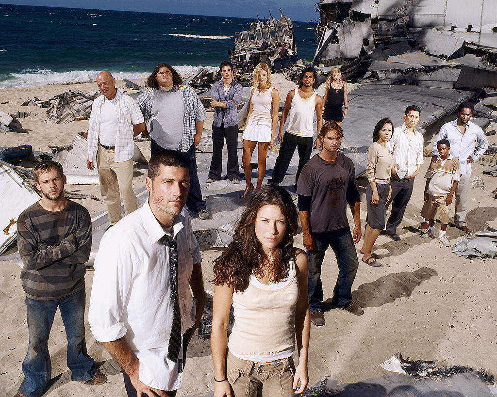 large cast standing on a beach in front of a wreckage