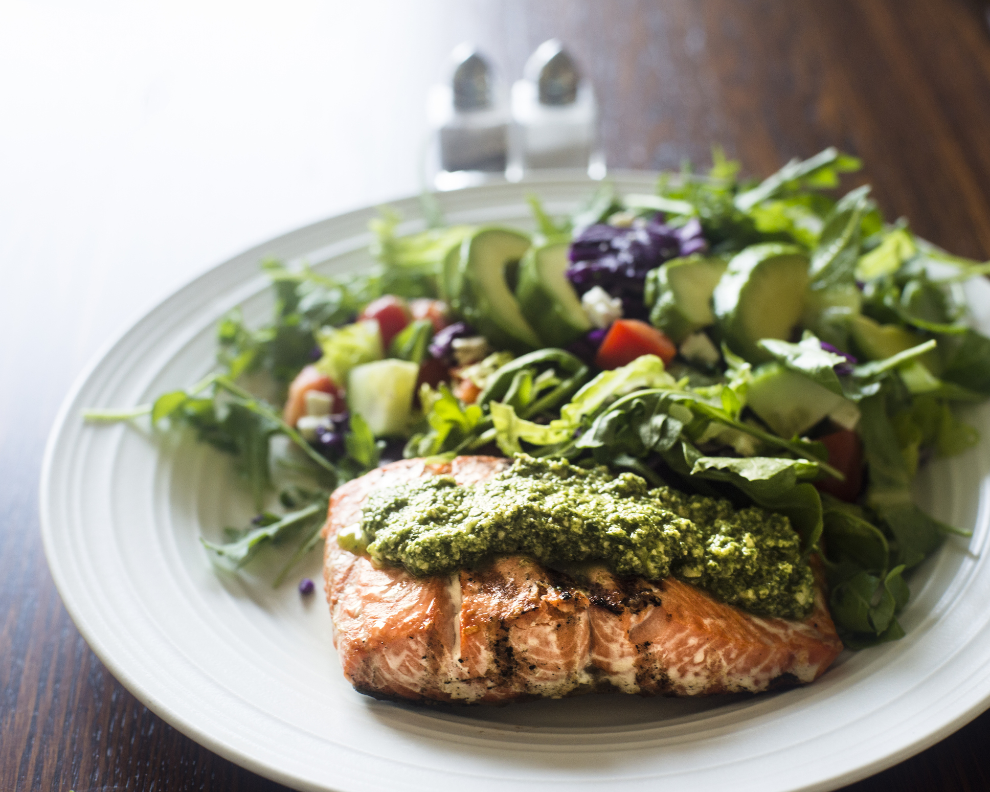 Close up of a healthy home-cooked meal on a plate which includes a grilled salmon fillet topped with arugula pesto accompanied by a green salad with avocado, romaine lettuce, cucumber, tomatoes, carrots and red cabbage