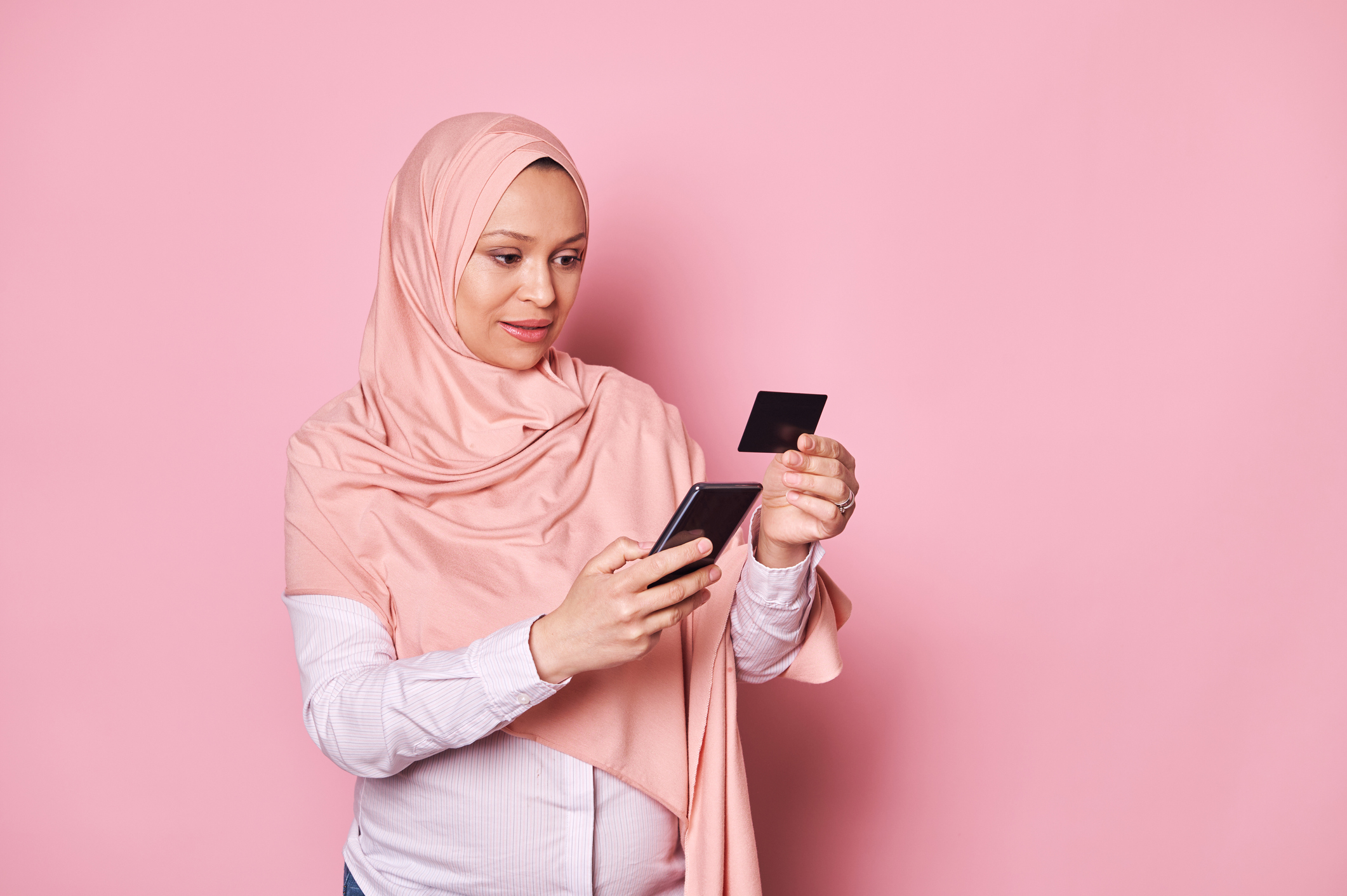 Happy pregnant Arab Muslim woman in hijab, holding credit card and smartphone