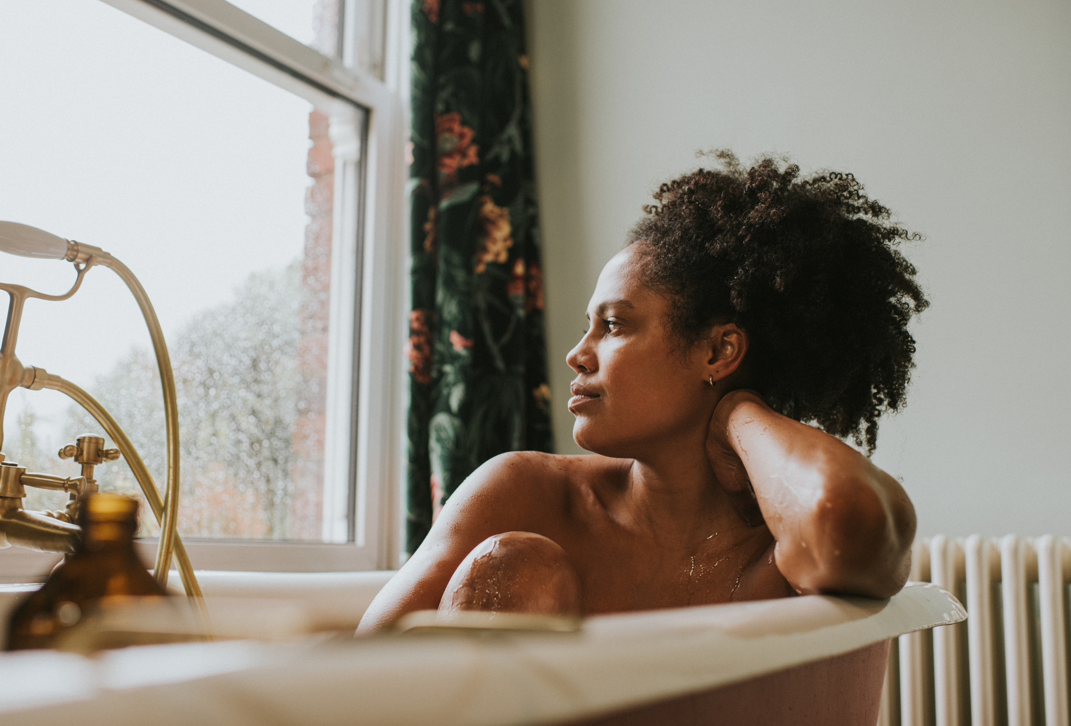 A Black woman bathes in a roll top bath, she seems distracted as she rests her head on her arm and gazes out the window