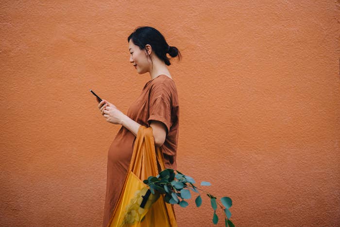 Young Asian pregnant woman carrying a yellow reusable shopping bag and using smartphone, shopping for fresh groceries in the city