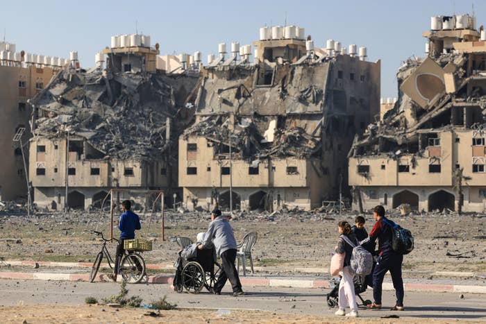 displaced palestinians walking with their belongings with bombed houses behind them