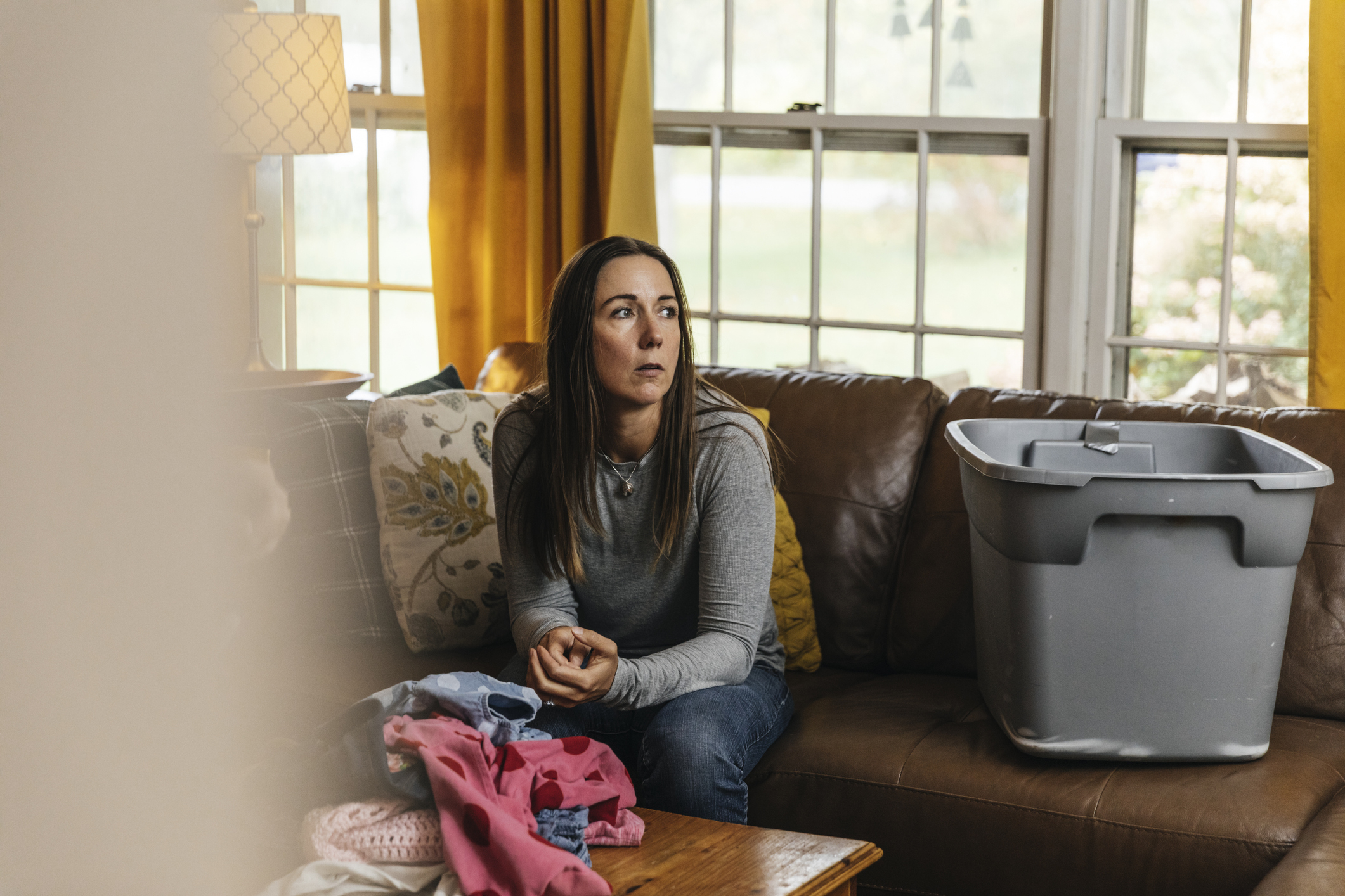 A woman is sitting in the living room next to a pile of clothes looking distraught