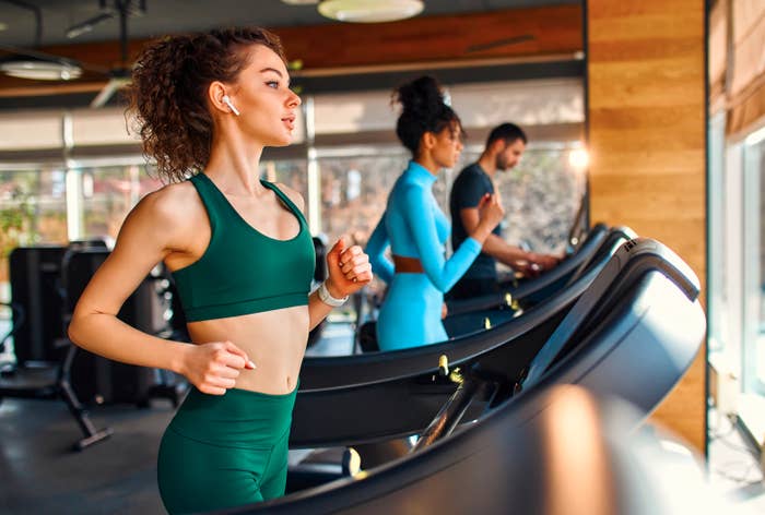a woman in a gym runs on a treadmill with her airpods in while another woman and a man also use treadmills in the background