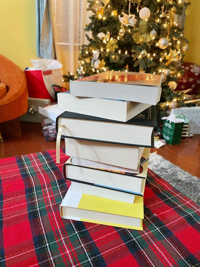 A stack of books are on a plaid table covering with a Christmas tree in the background