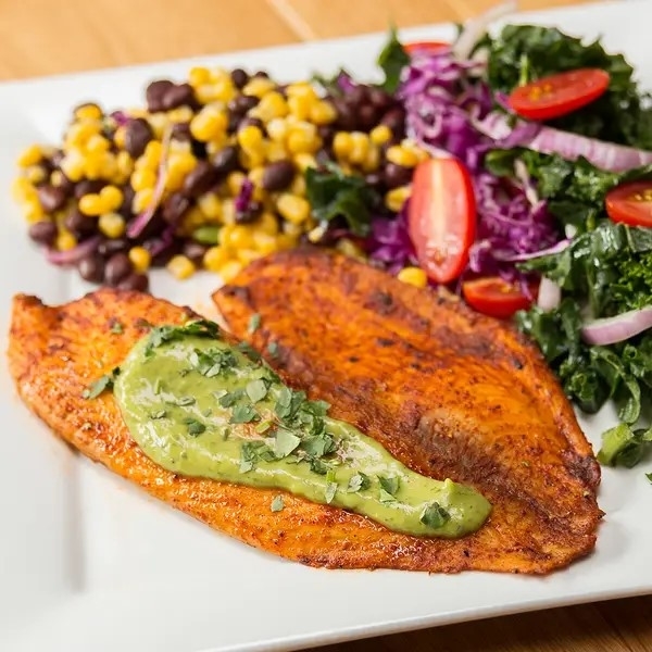 Grilled fish fillet with avocado sauce, served with a side of corn and black beans, and a mixed salad of kale, red cabbage, cherry tomatoes, and red onion