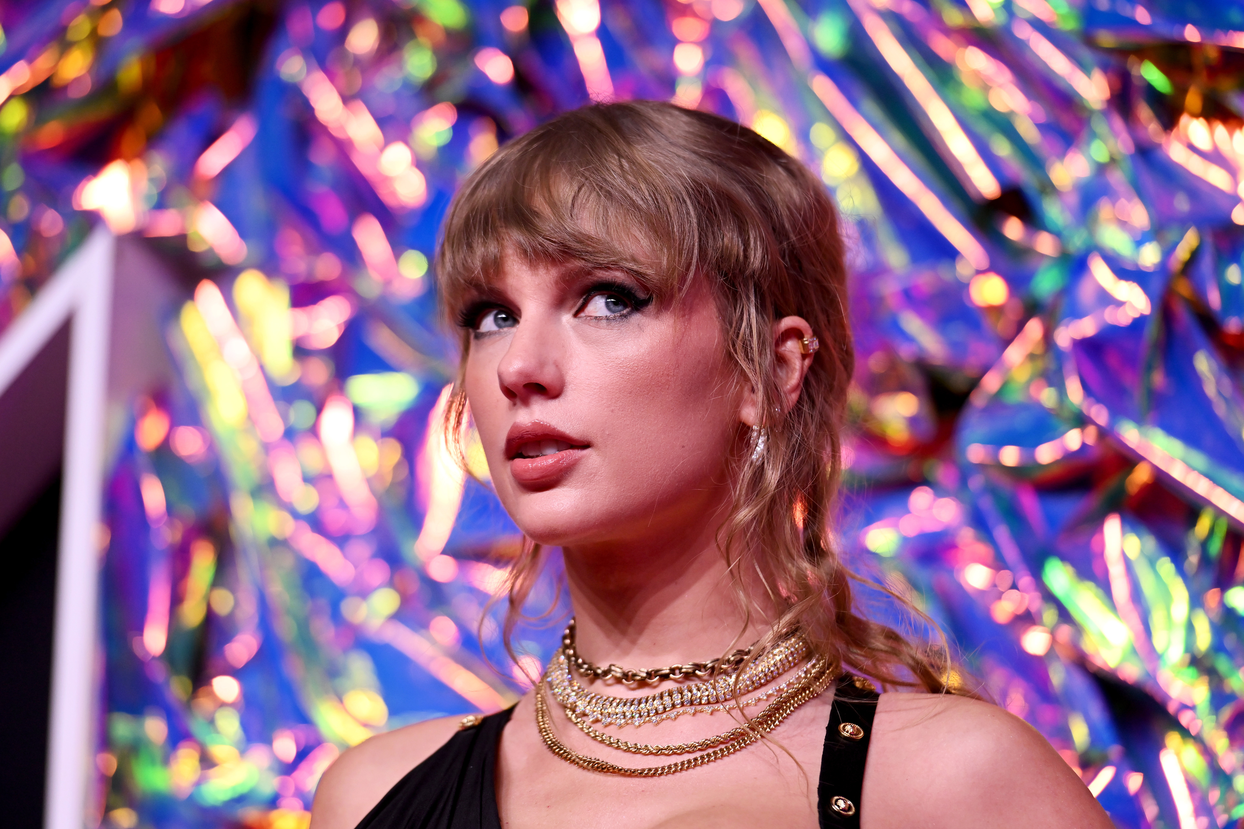 Close-up of Taylor wearing many necklaces and multicolored lights behind her