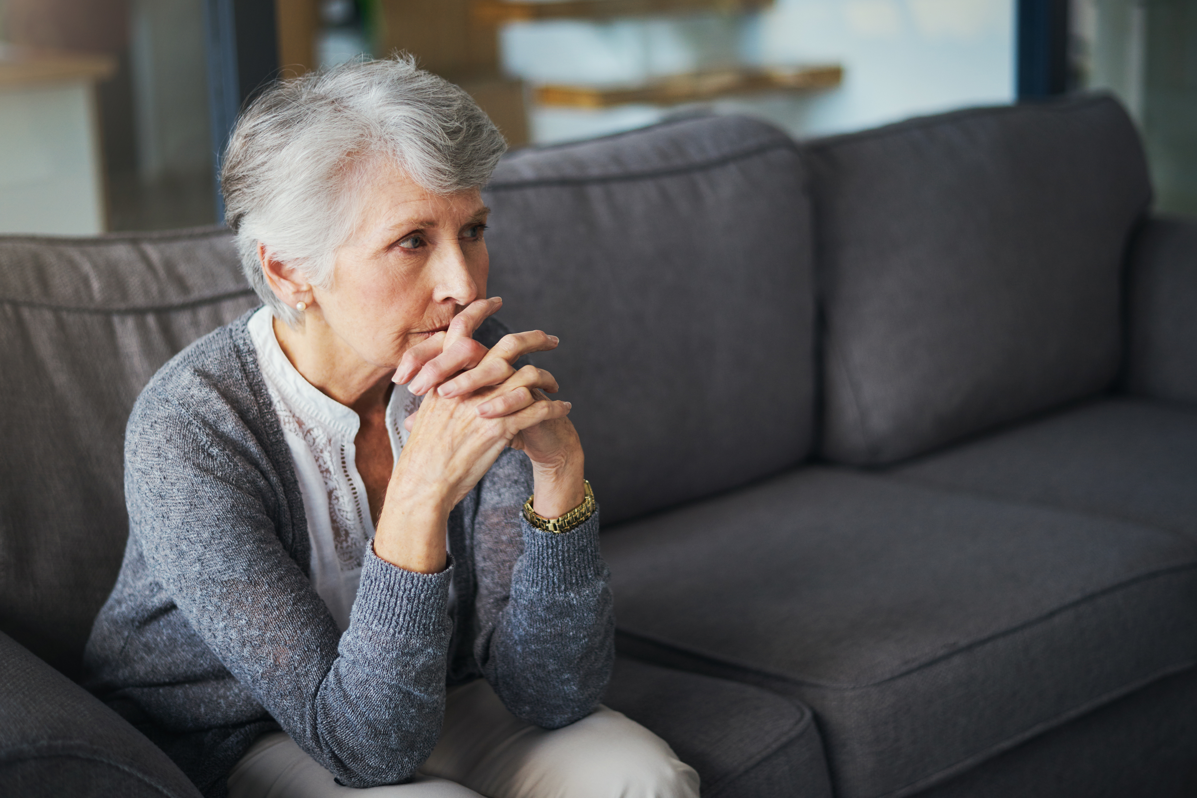 An older woman sitting on a couch with her hands clasped and her elbows resting on her knees