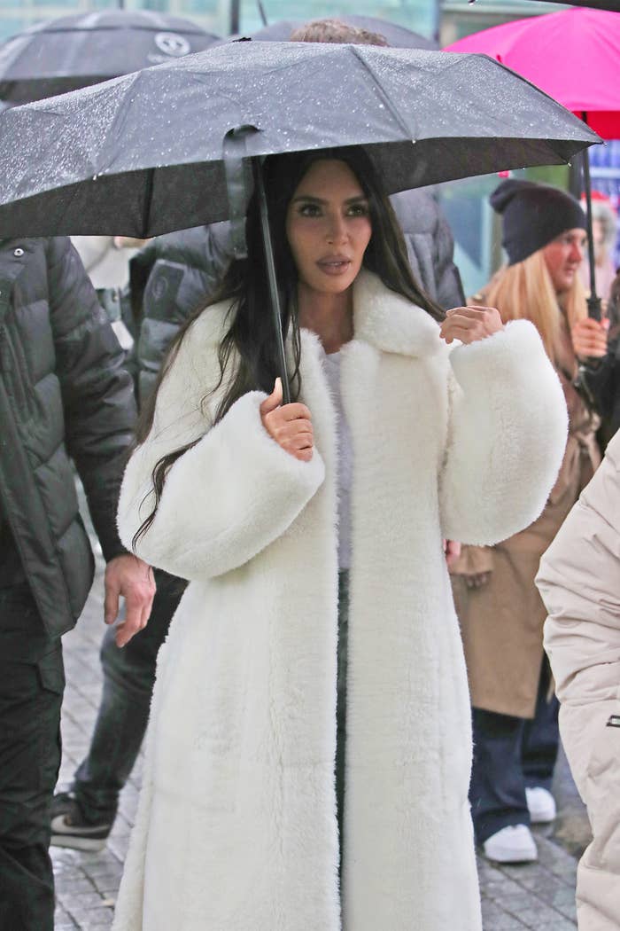 Kim Kardashian in a long, furry white coat and holding an umbrella