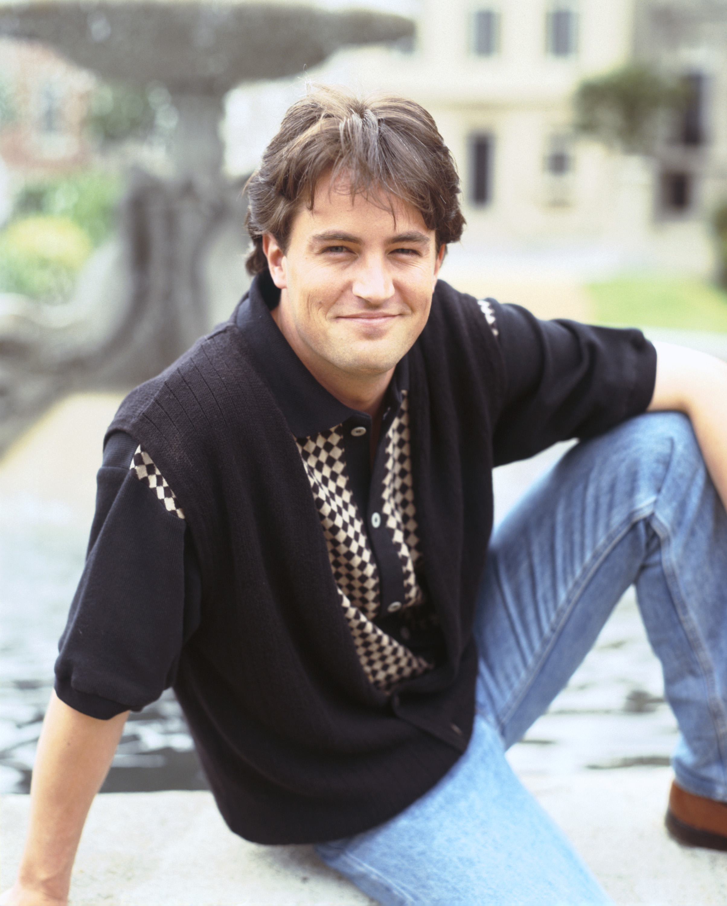 Close-up of a younger, smiling Matthew sitting down and wearing jeans, vest, and T-shirt