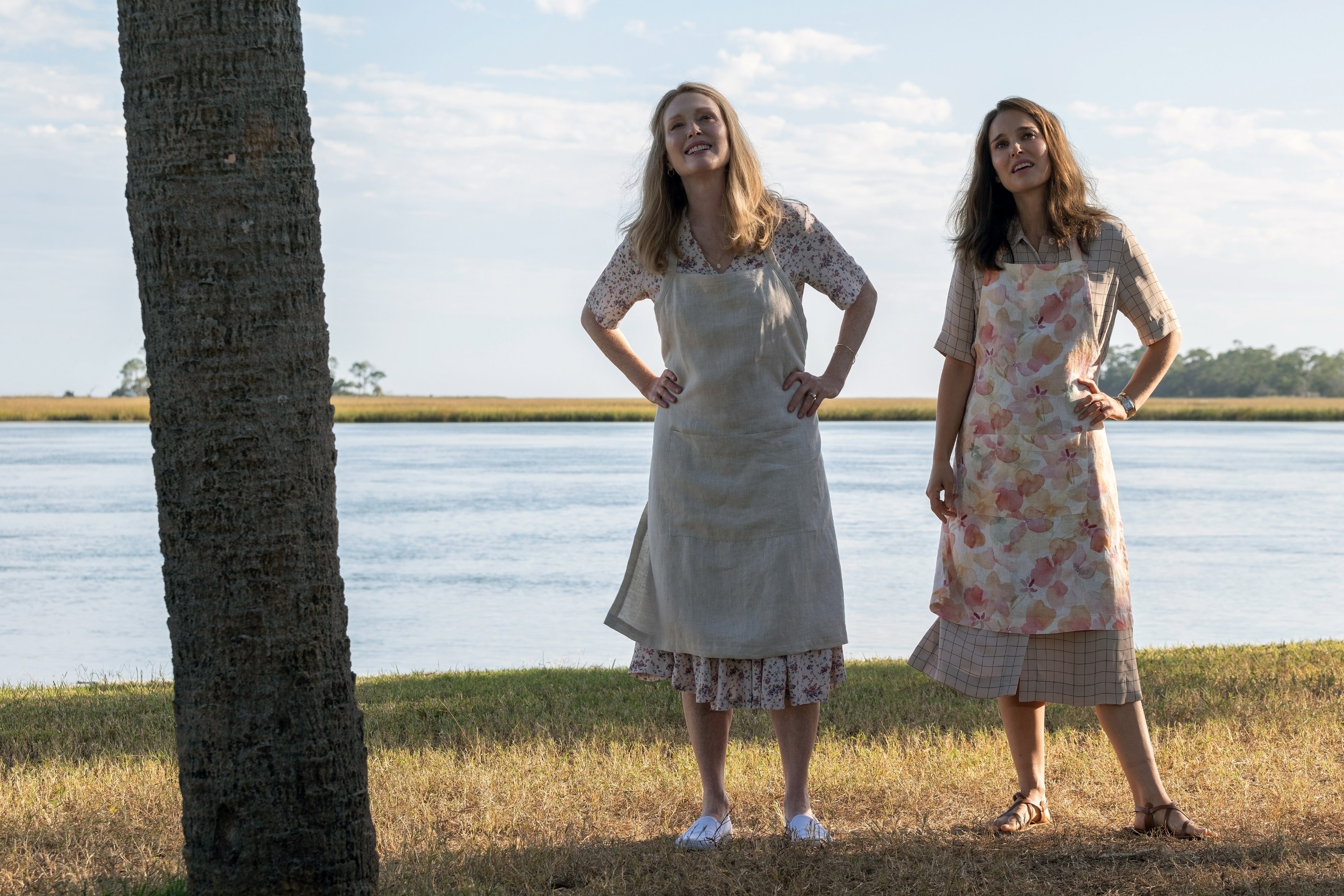 Julianne, on the left, standing next to Natalie with a body of water behind them in a scene from &quot;May December&quot;. They are both wearing aprons