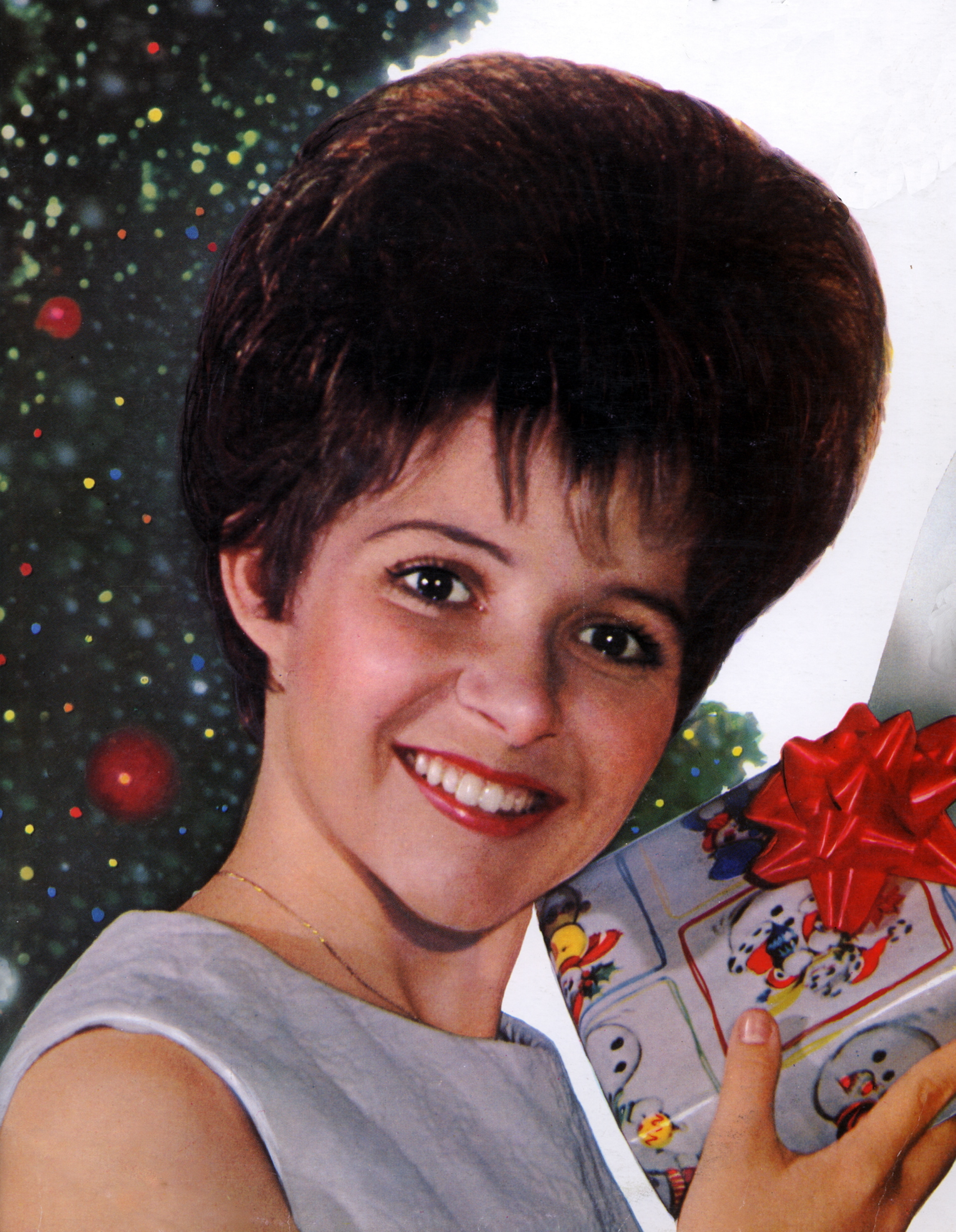 Close-up of a young Brenda smiling in front of a Christmas tree and holding a wrapped gift