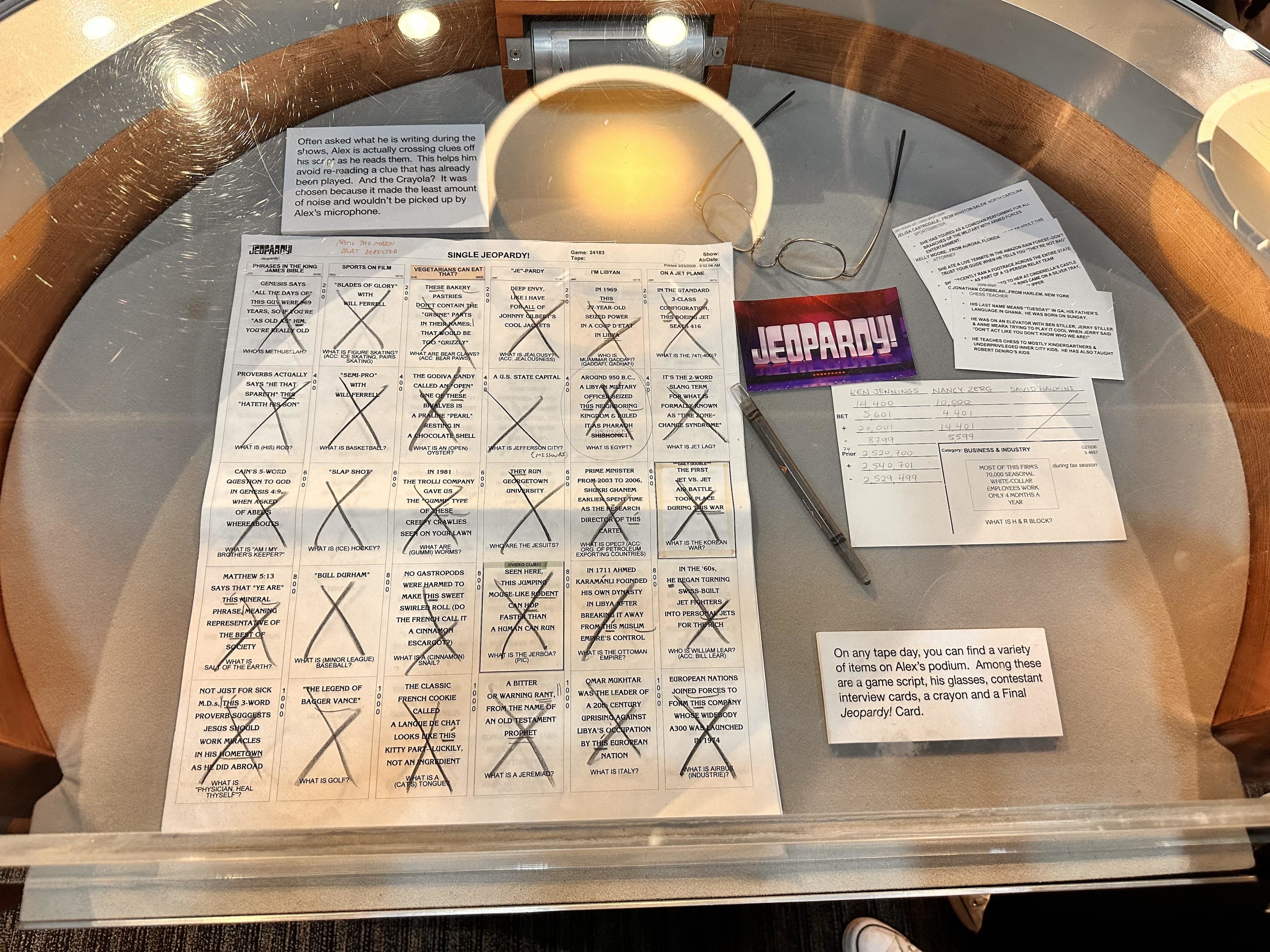 A podium showing a sheet of paper with clues X-ed out, contestant interview cards,  eyeglasses, a crayon, and a Final Jeopardy card