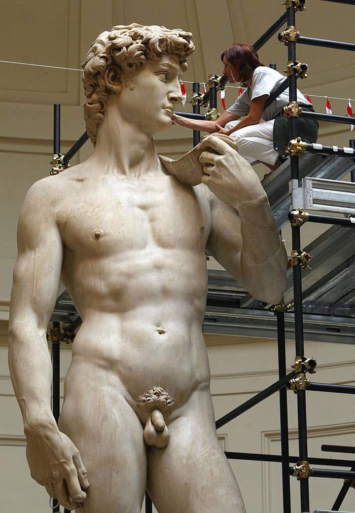 Close-up of the nude sculpture, with a person on scaffolding at the top, crouched and working on the head, who looks no bigger than the head