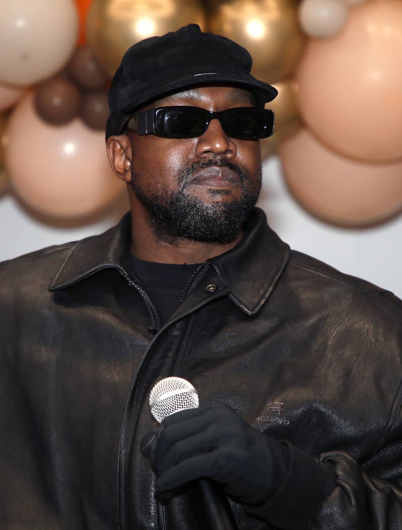 Close-up of Ye in leather, a cap, and sunglasses and holding a microphone