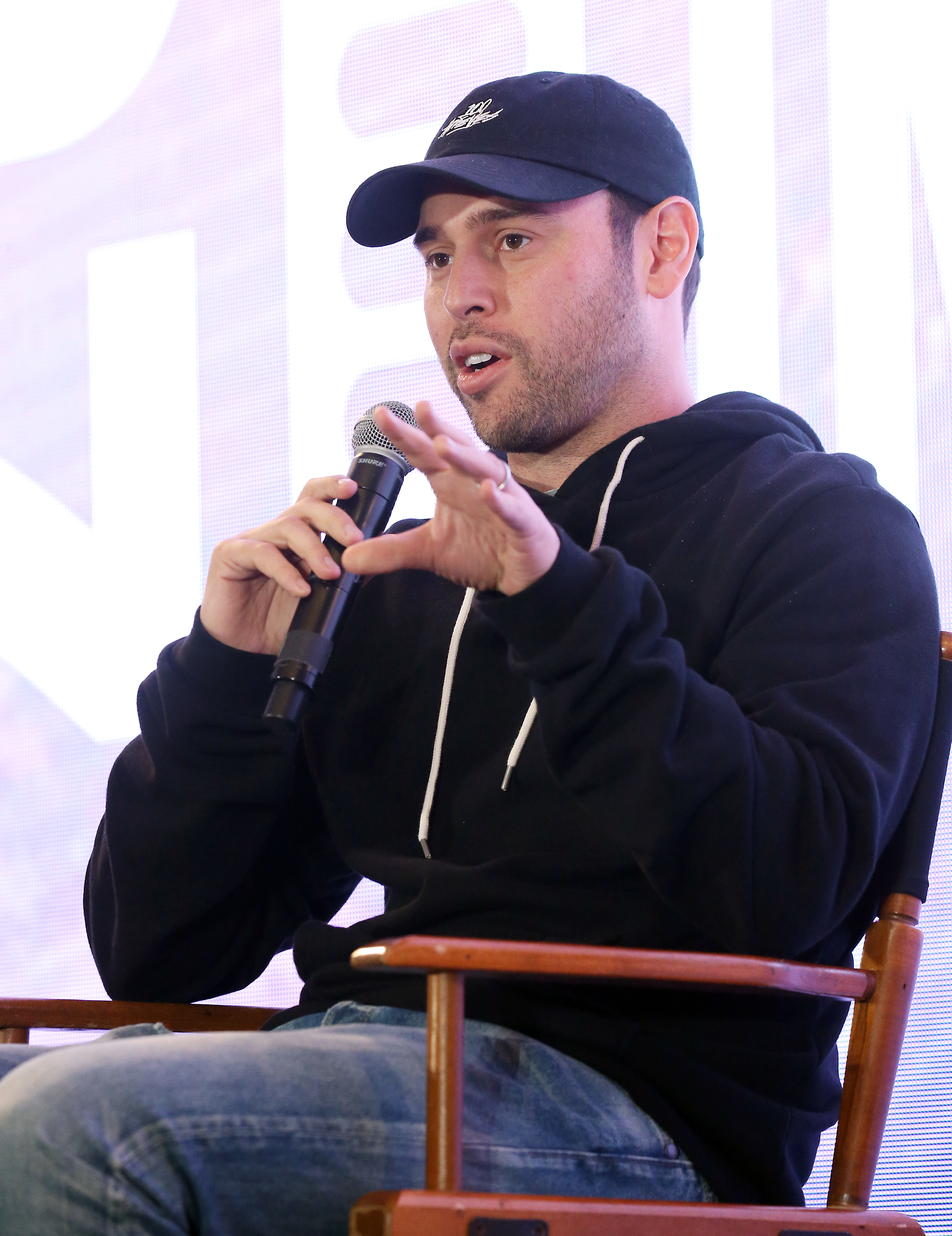 Close-up of Scooter sitting onstage with a microphone and wearing a cap, hoodie, and jeans