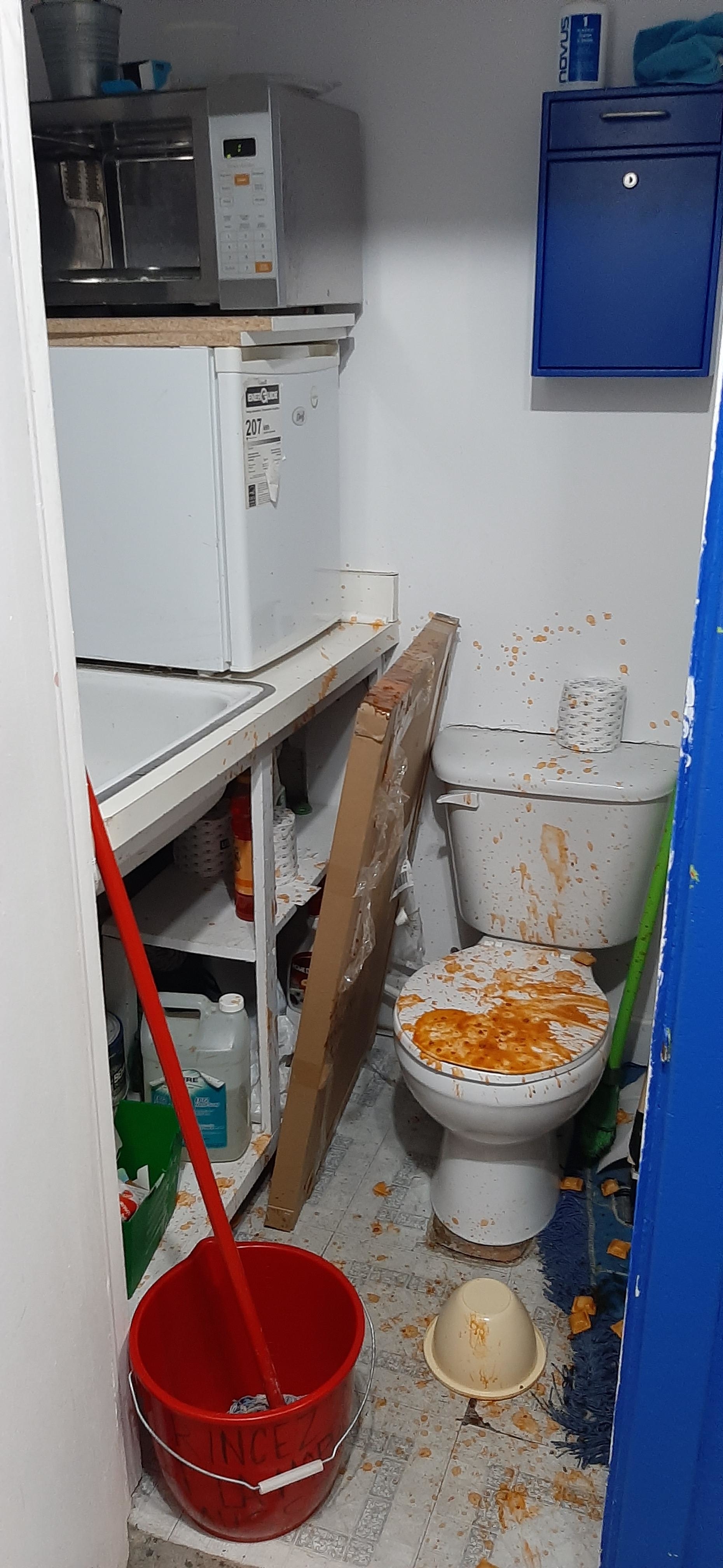 A small room that has a mini fridge and microwave stacked on a counter. There is a toilet next to the counter that is splattered with ravioli sauce