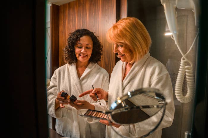 Two mature women in white hotel robes are getting ready in their hotel bathroom