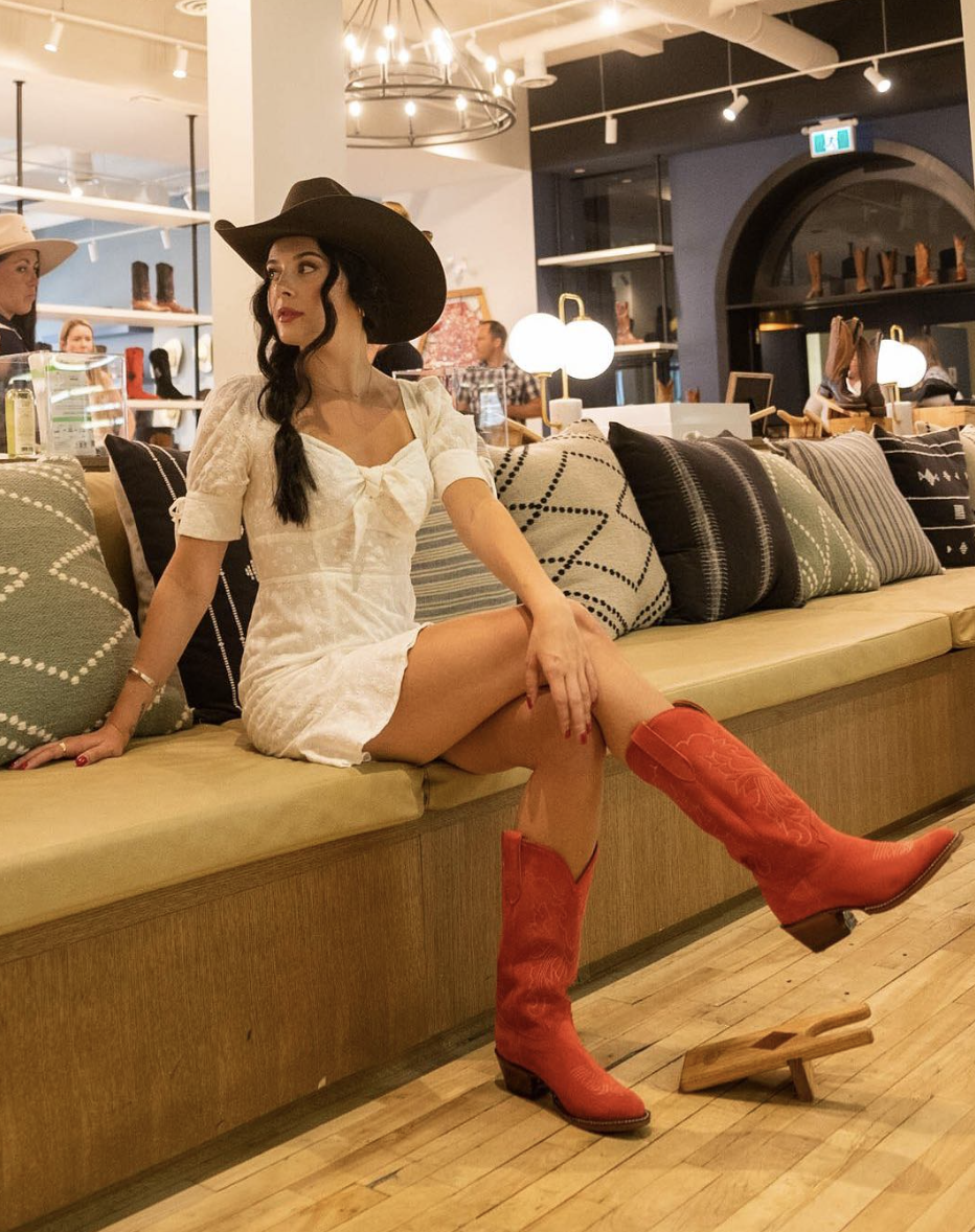 A woman sits on a couch and wears cowboy boots and a cowboy hat.