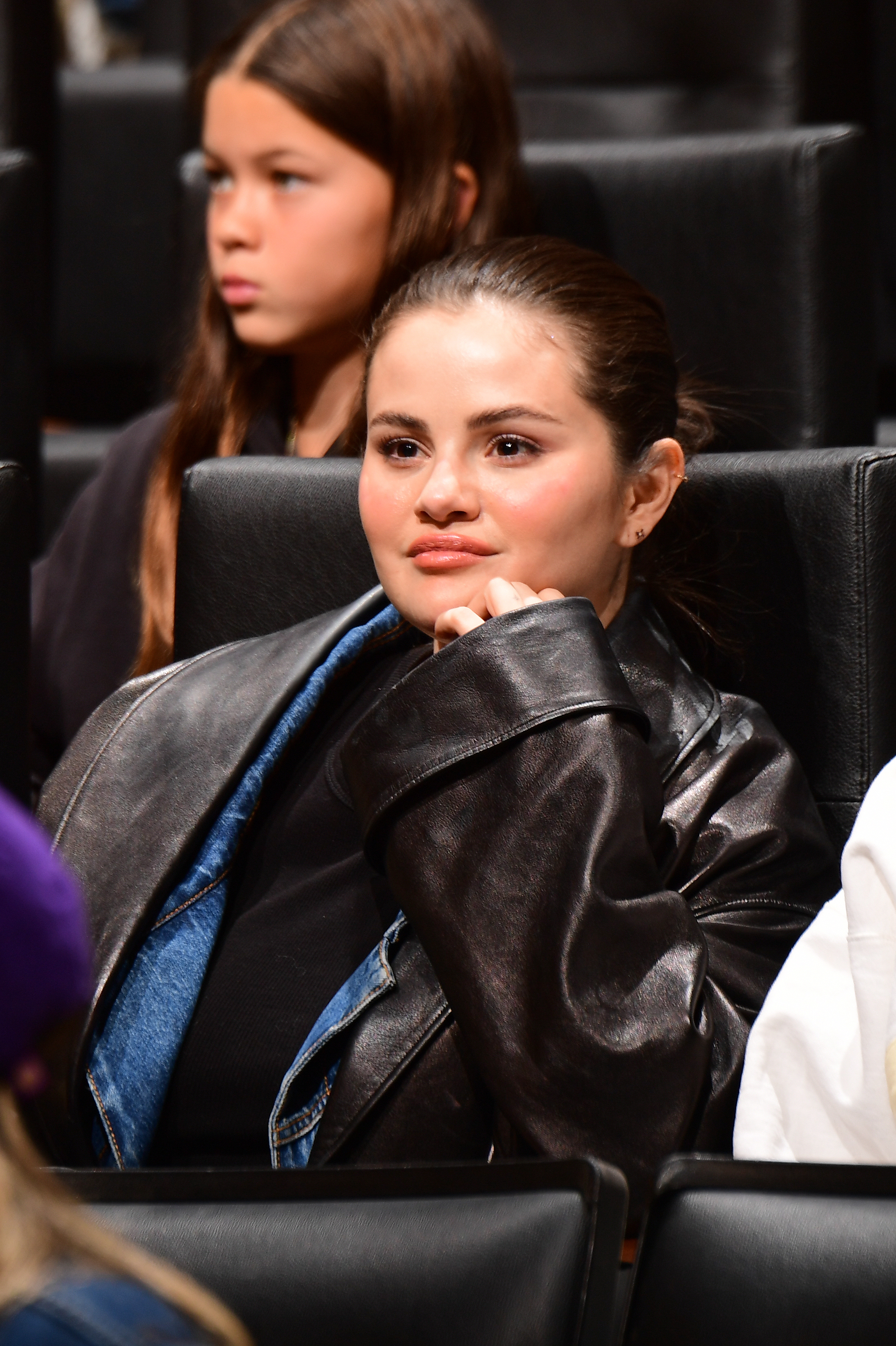 Close-up of Selena seated in an audience, smiling slightly, and wearing a leather jacket