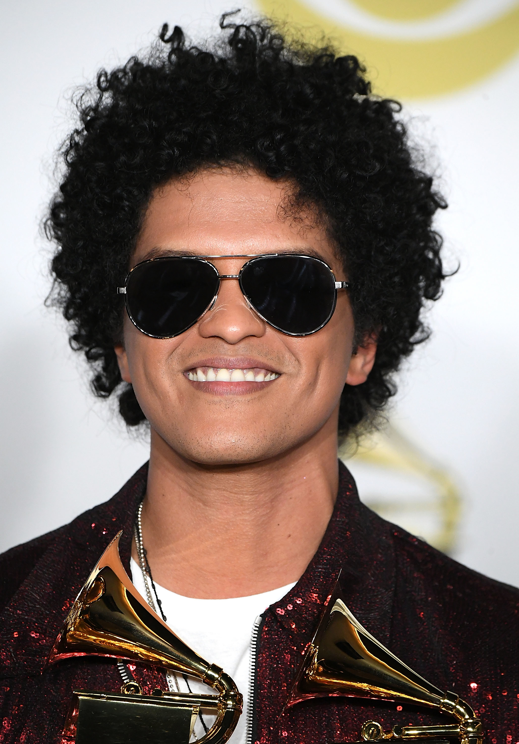 A closeup of Bruno Mars wearing sunglass and smiling as he holds two Grammys