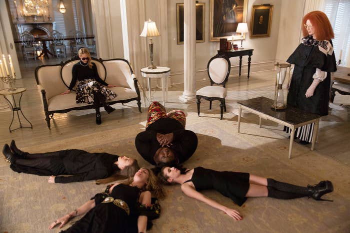 A group of girls lay in a circle on the ground as two other witches look on.
