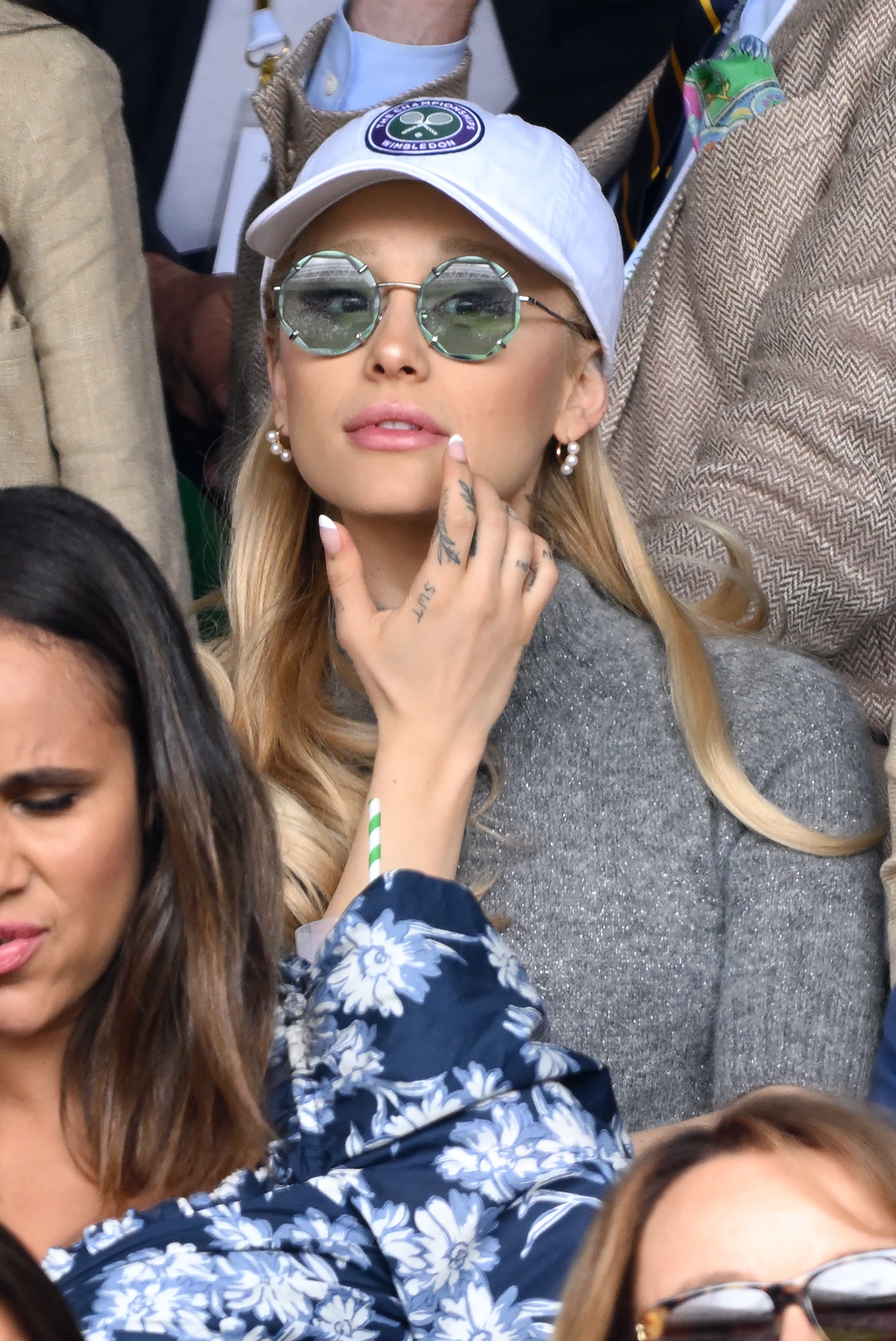 Close-up of Adriana sitting in an audience wearing a cap and sunglasses