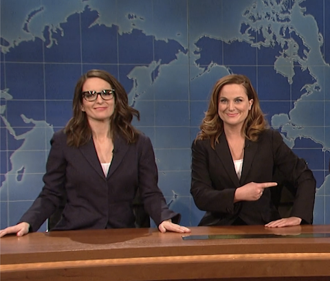 tina fey and amy poehler sitting at a desk with a world map behind them