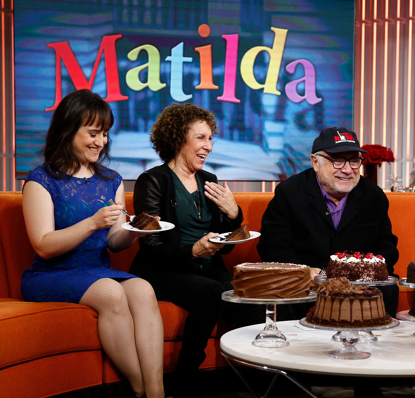 Mara, Rhea, and Danny eating cake on a talk show