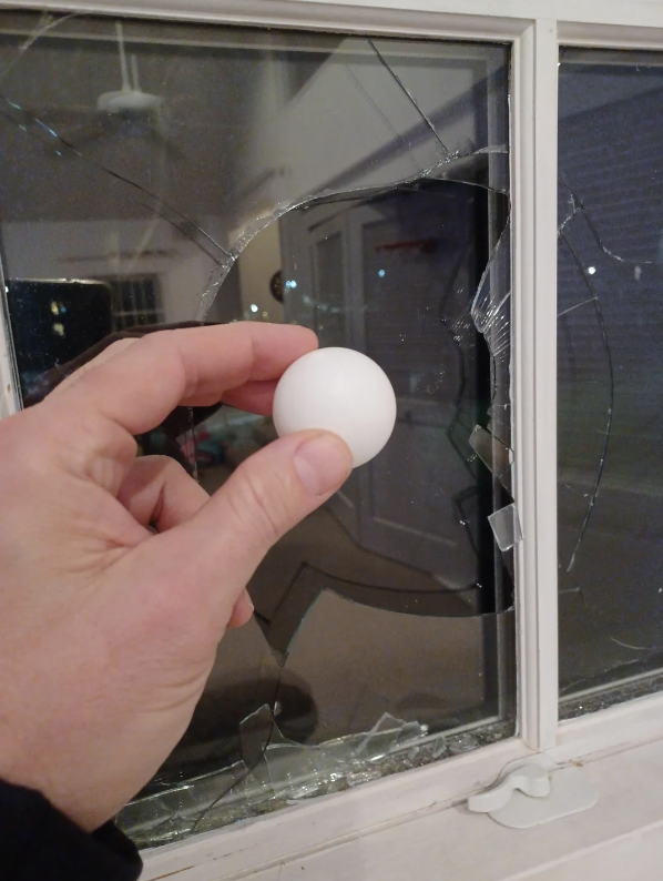 a hand holding a ping pong ball in front of a broken window