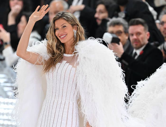 Gisele waving at a media event while wearing a sparkled dress with a feathered cape