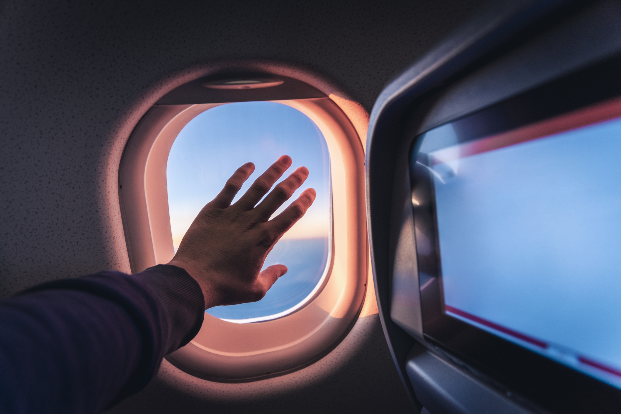 Personal perspective of human hand touching the airplane&#x27;s window while flying above the sea at sunrise