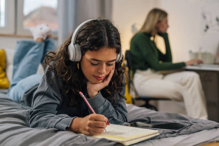 Two young women in a room, one on the bed with headphones and the other sitting at a desk