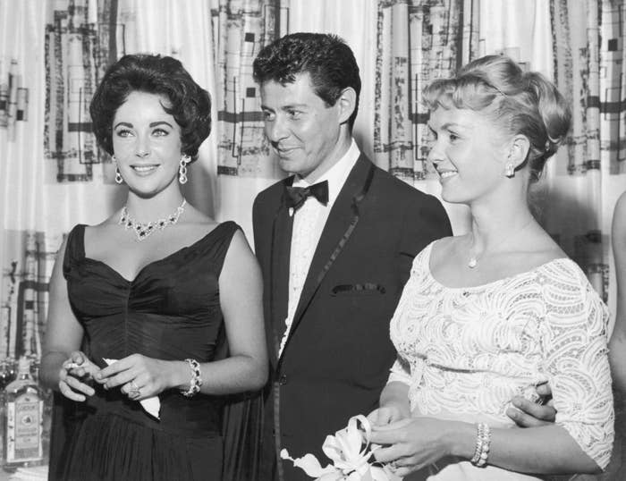American singer Eddie Fisher, wearing a tuxedo, stands with arm around his wife, American actor Debbie Reynolds (R) and smiles while looking at British-born actor Elizabeth Taylor