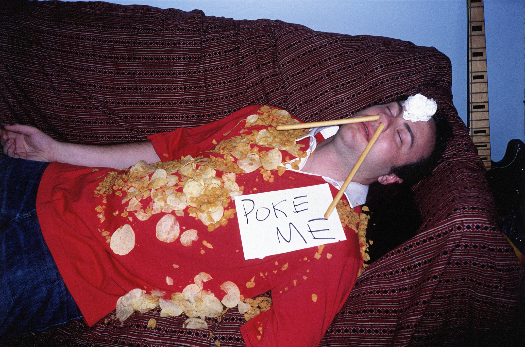 A very drunk person lying on a couch with potato chips all over their T-shirt, breadsticks emerging form their nostrils, and a &quot;Poke Me&quot; sign on their chest