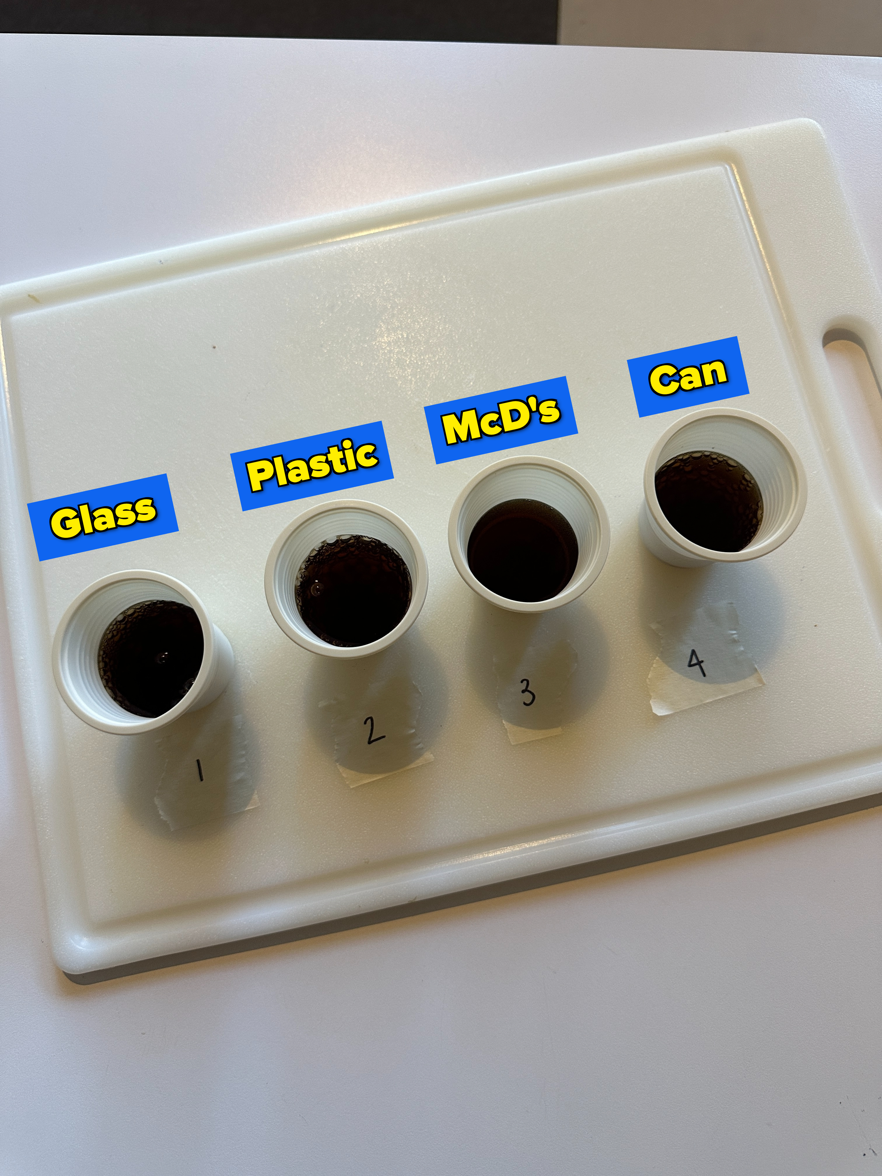 Four mouthwash cups on a cutting board, each filled with Diet Coke from different vessels