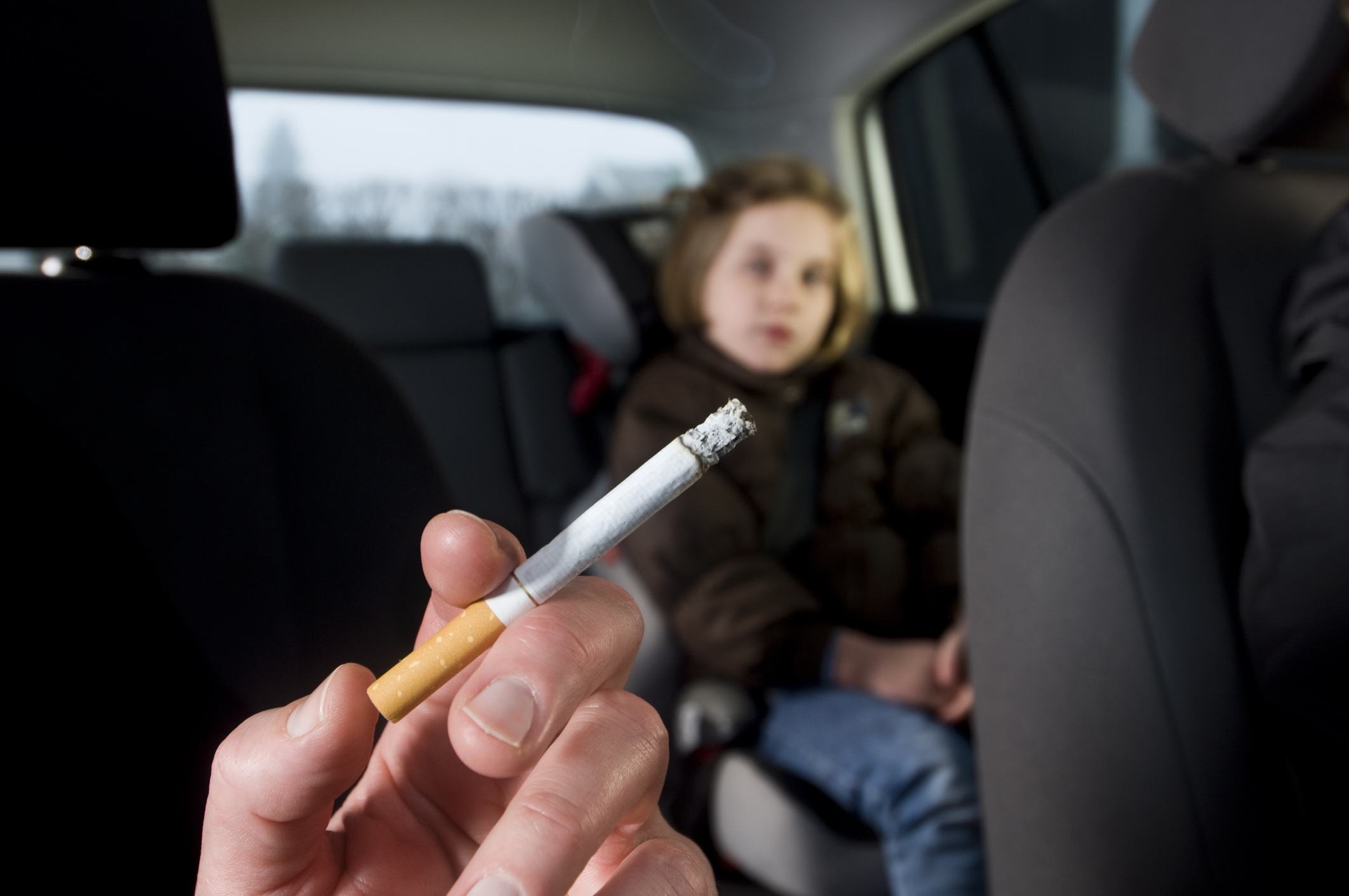 A parent is smoking a cigarette in a car with their kid in it