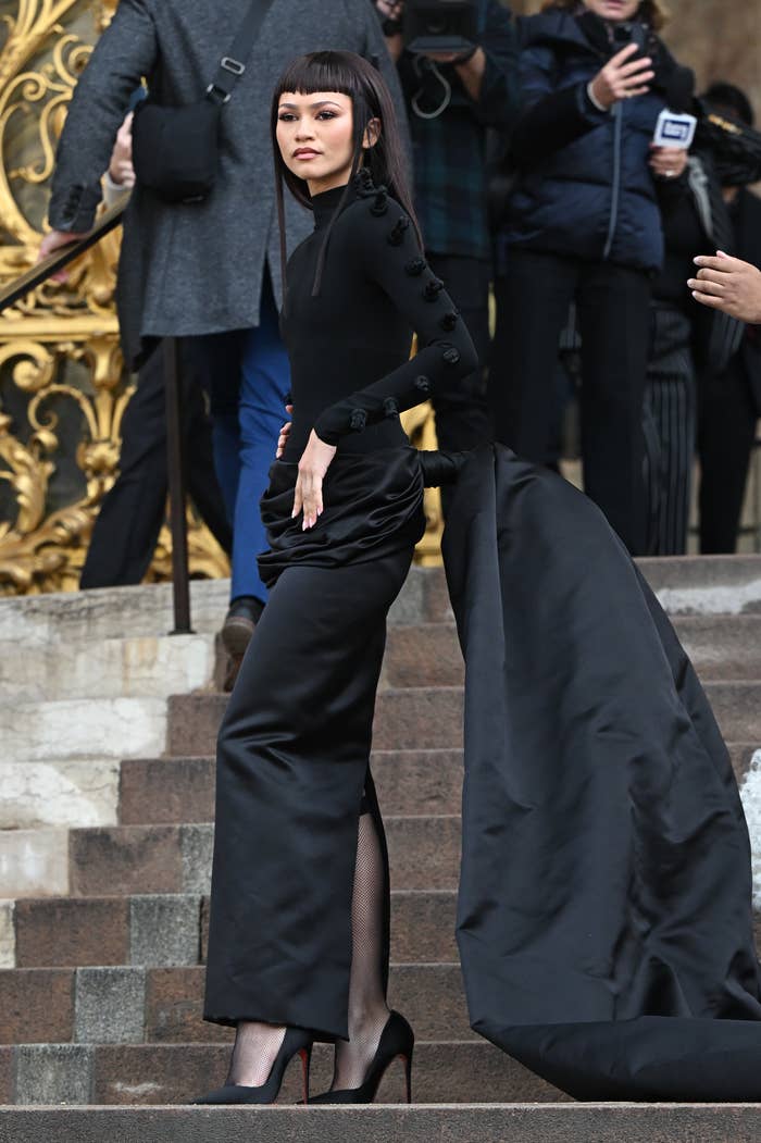 Zendaya standing on the steps outside the show in a black turtleneck, long-sleeved straight gown with back slit