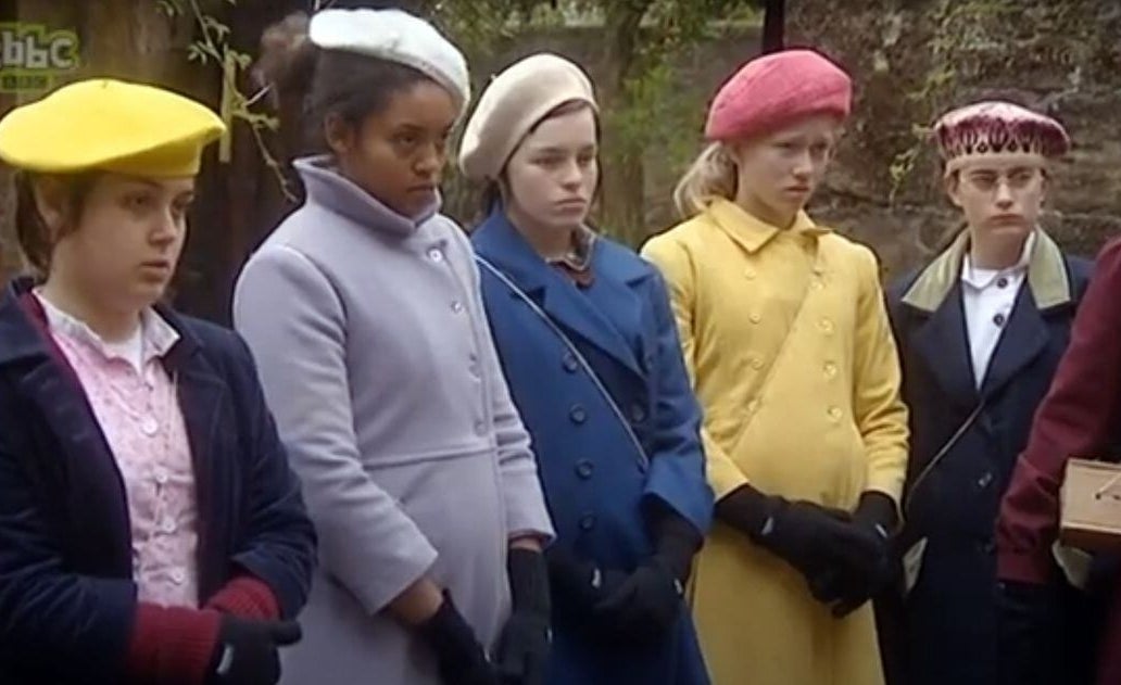 Five girls stand in a line wearing 1940s style clothing with gas mask boxes across their shoulders.