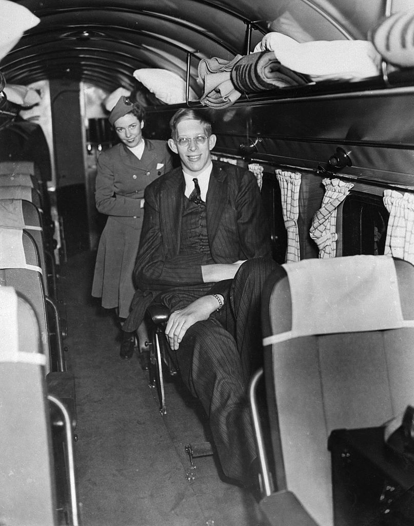He&#x27;s wearing a suit and sitting with one leg stretched out on an old, single-row plane and a flight attendant behind him
