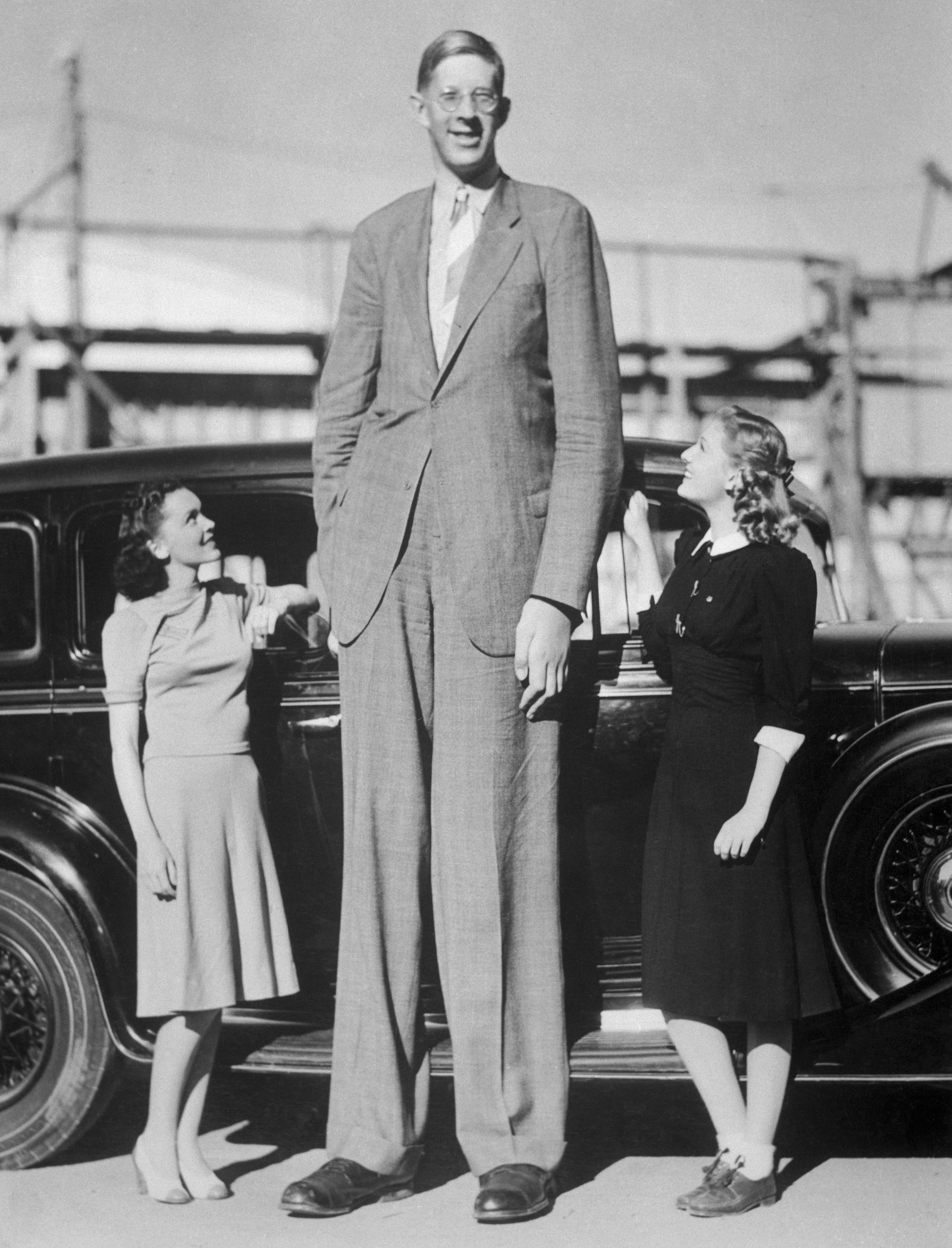 He&#x27;s wearing a suit and standing in front of a car next to two smiling women, one of whom comes to his hip and another to his waist