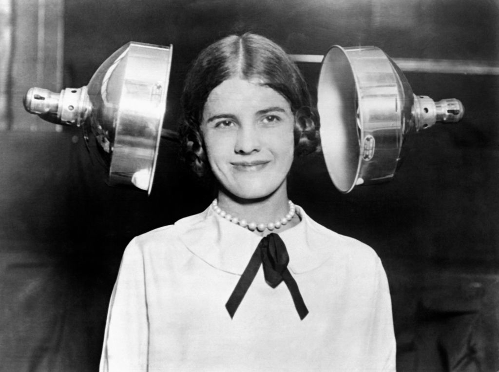 A smiling woman sits between two large metallic, round blowers that look as if they&#x27;re about to squeeze the sides of her head