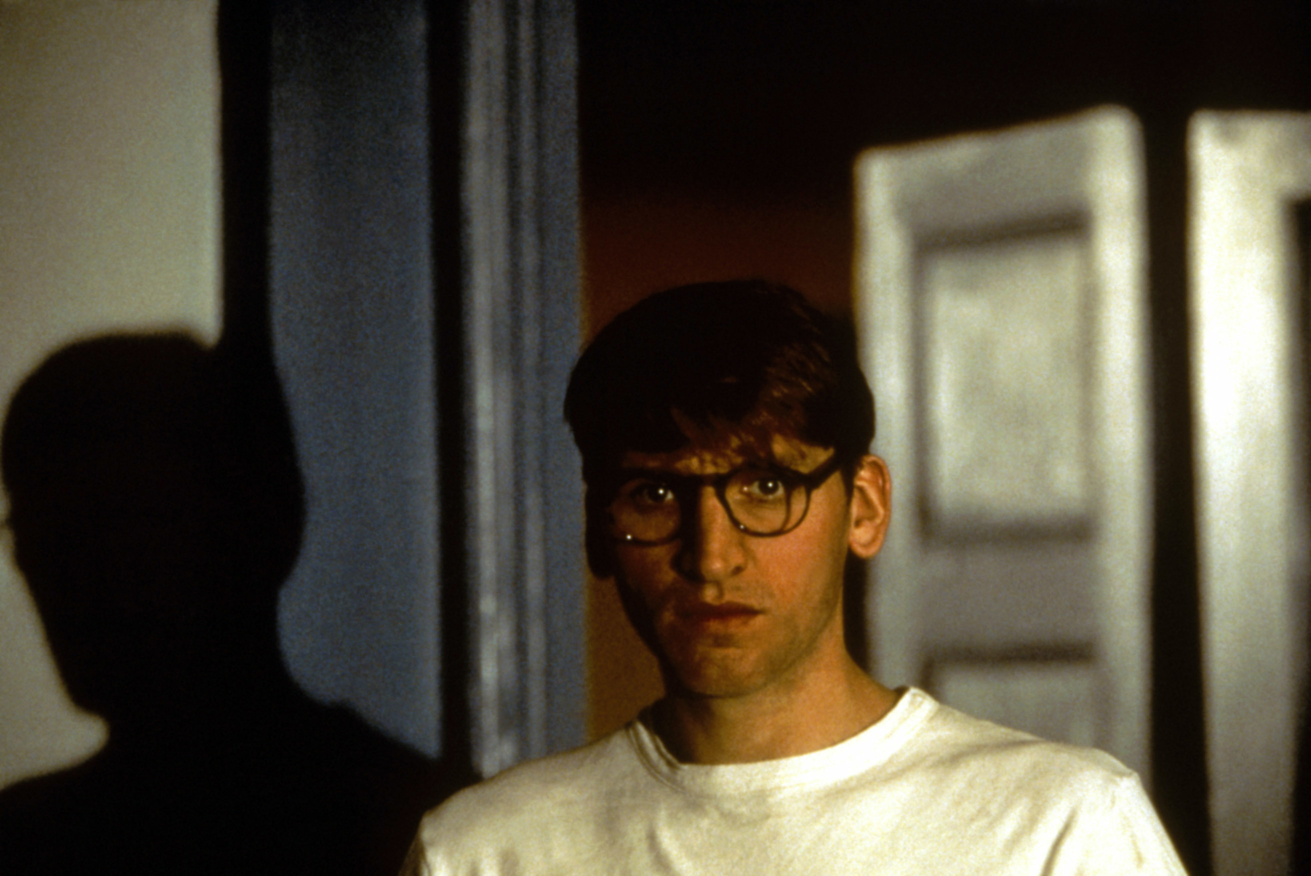 A young man with brown hair and thick glasses, looks out suspiciously a doorway.