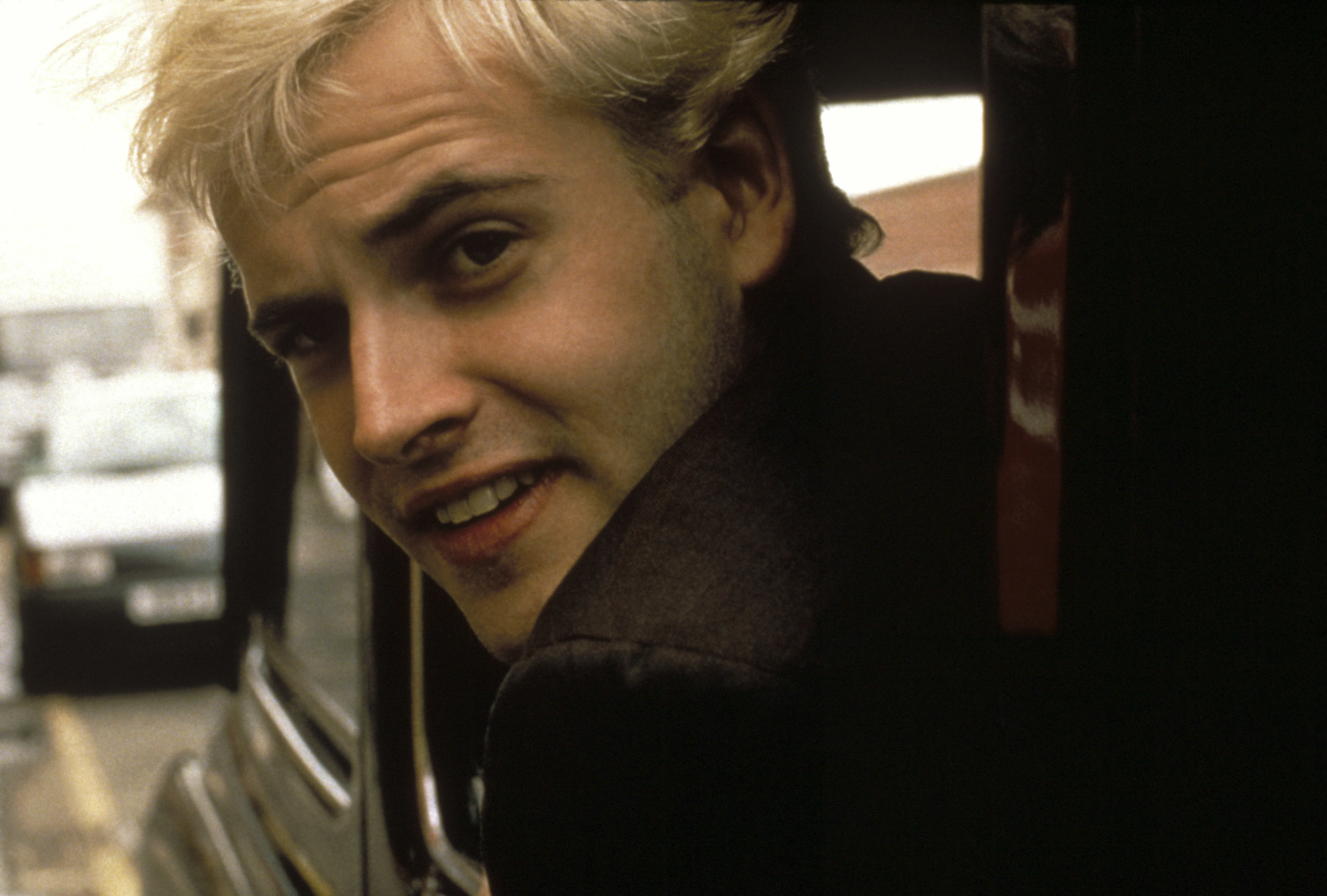 Young man with scruffy bleached blonde hair, looks out of a car window over his shoulder.
