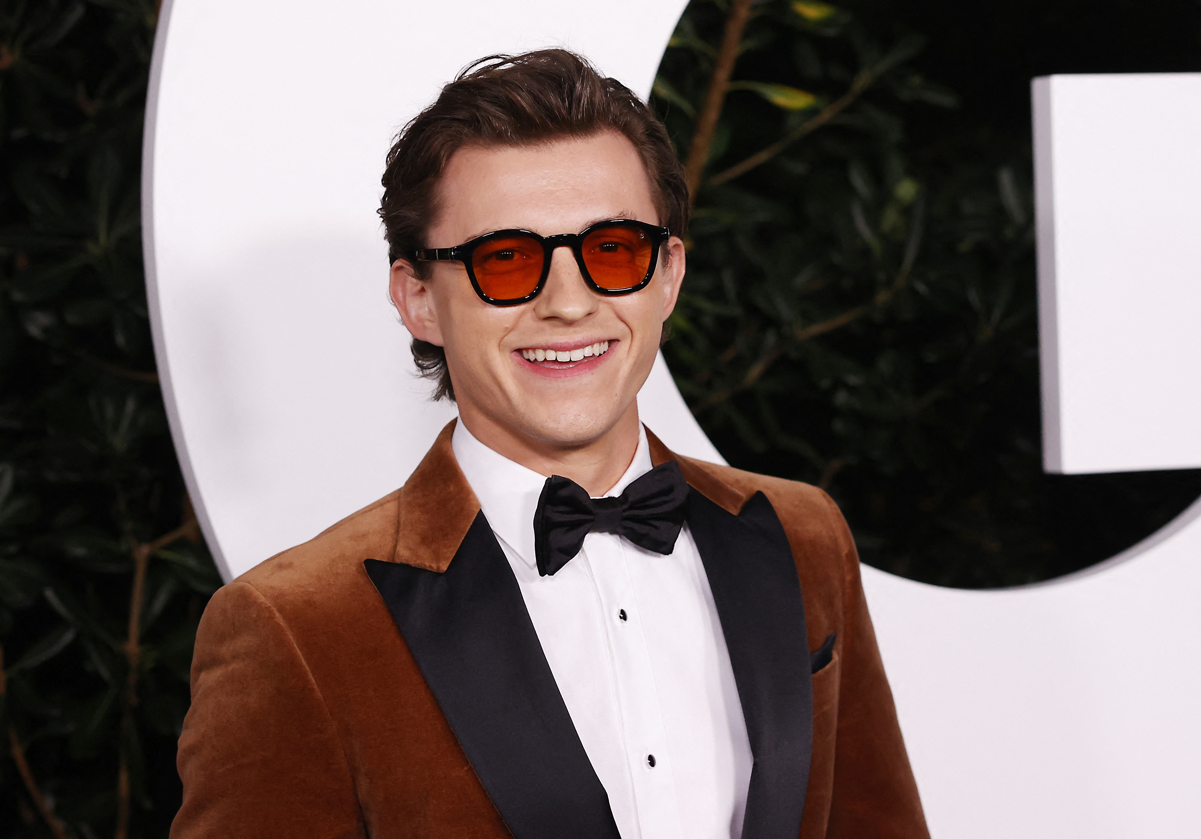 Close-up of Tom Holland smiling in sunglasses and a suit and bow tie at a media event