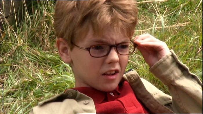 A boy lays on grass and fixes his glasses with a confused look.