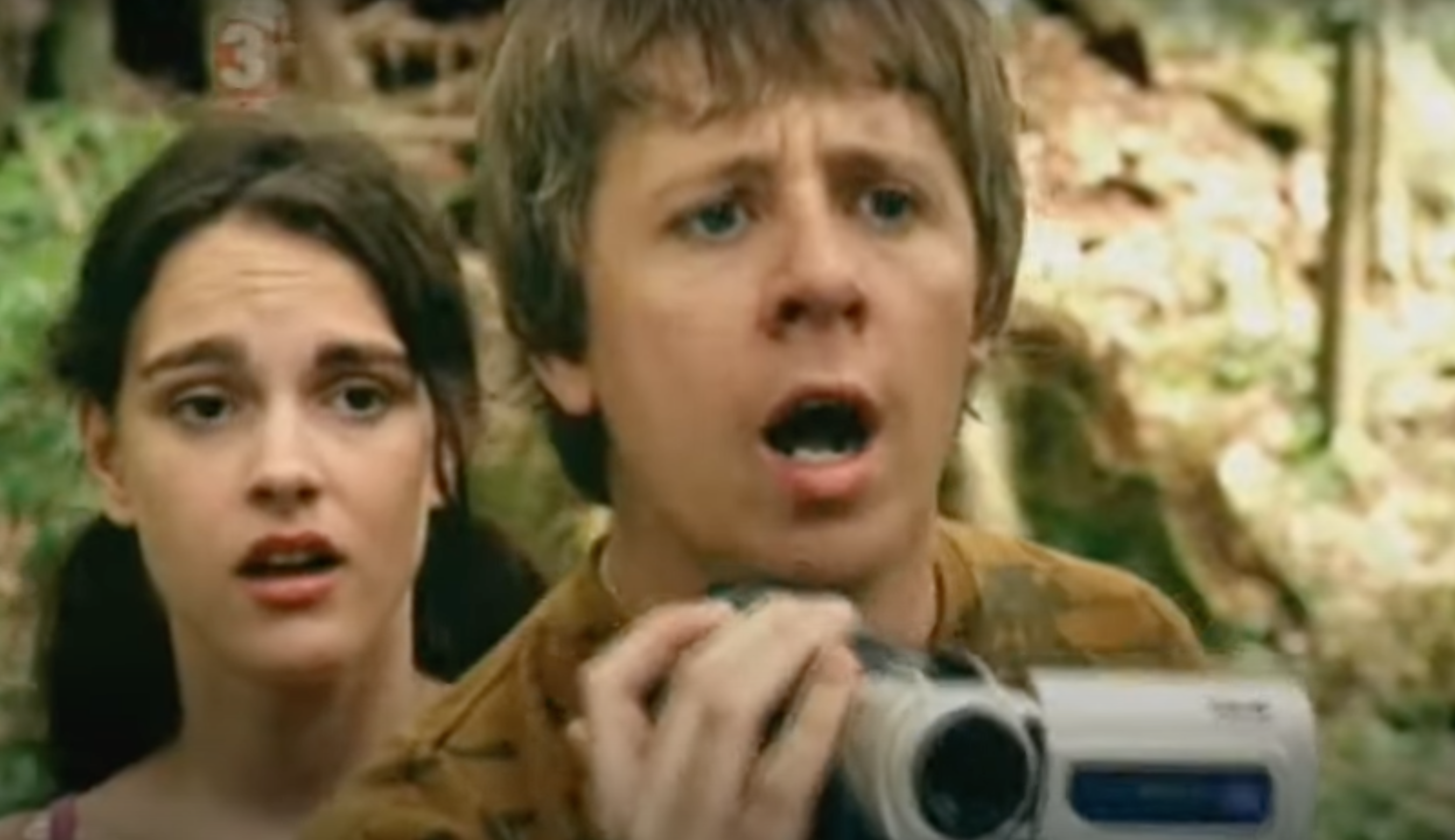 A boy holds a camcorder in shock as a girl stands behind him in the woods.
